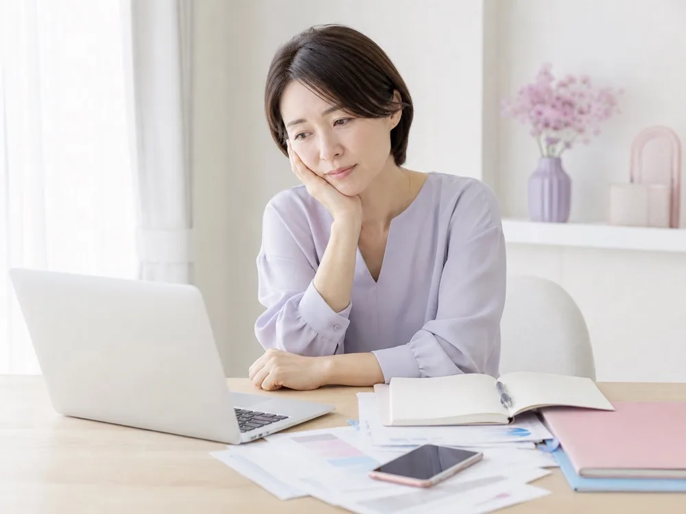 ノートパソコンと書類に囲まれ、ひとりで仕事を抱えて考え込んでいる女性
